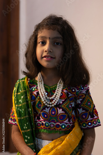 Portrait of a young Indian girl wearing traditional Gujarati ethnic clothing with a green patterned dupatta, colorful embroidered blouse, and layered pearl necklace.