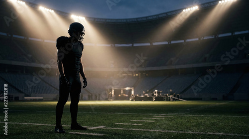 American football player in full protective gear standing on green turf under bright floodlights in dramatic empty stadium at night