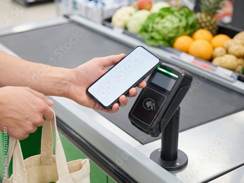 A young adult customer uses a smartphone for contactless payment at a grocery store checkout counter while holding a reusable fabric shopping bag
