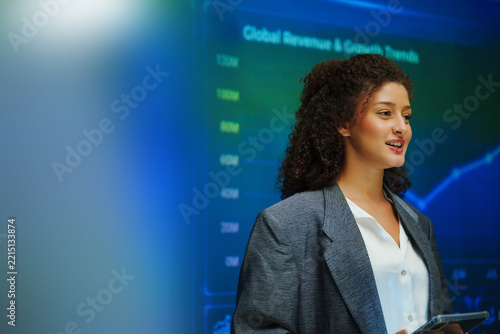 Hispanic Woman gives a business presentation in front of a digital dashboard with charts and graphs, highlighting data analytics, performance and strategic insights.