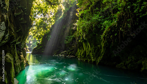 Sunbeams illuminate a vibrant green river flowing through a lush mossy canyon with dense foliage overhead