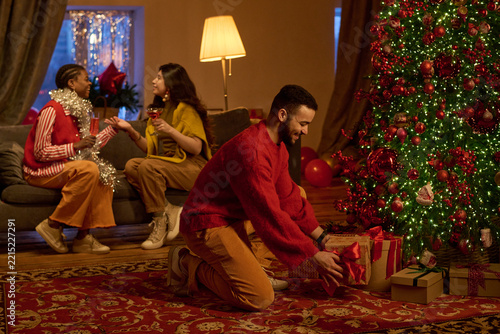 Young adult man placing gift box under decorated Christmas tree near multiethnic friends celebrating winter holidays. Ideal for seasonal advertising and festive retail campaigns