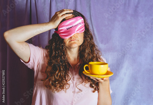 Tired woman holds a cup of tea while wearing a pink sleep mask