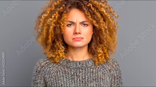 A frustrated woman with curly hair covering her ears and yelling against a grey background.