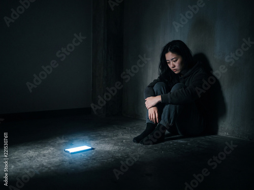 Young woman sitting alone in dark corner hugging knees, glowing smartphone on concrete floor, digital fatigue, anxiety and loneliness concept