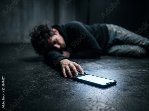 Exhausted man lying on dark concrete floor reaching for glowing smartphone, digital fatigue and phone addiction concept