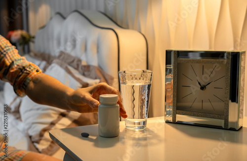 Unrecognizable woman reaching hand for pill bottle on bedside table at night. Cozy bedroom interior with warm evening light, Insomnia concept and sleep hygiene