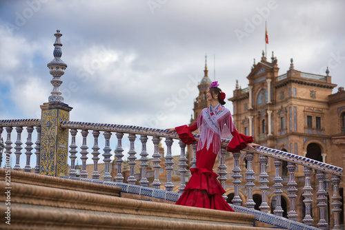 A young Andalusian woman wearing a red flamenco dress with ruffles and a purple shawl, with flowers in her hair, posing while leaning against a beautiful bridge in Spain Square in Seville.