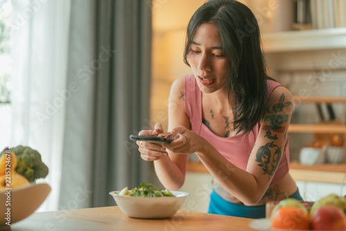 Young Asian woman photographing bowl of fresh green salad with smartphone on kitchen table at home Asian woman photographing healthy salad with smartphone in kitchen