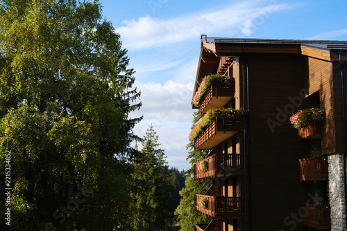 Obraz na plátně Side view of a traditional brown wooden alpine hotel facade with balconies decorated with colorful blooming flowers, next to green forest trees under a cloudy sky