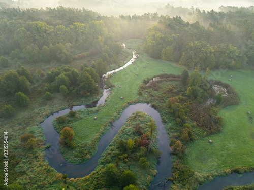 Aerial view of an S-shaped Rawka River meander winding through misty autumn woodland and meadows, Poland