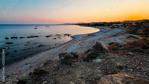 Serene twilight view of a rocky pebble beach curving along the coast of Rhodes, Greece, with calm sea reflecting a vibrant orange-to-blue sunset gradient, anchored boats, and a resort town visible in