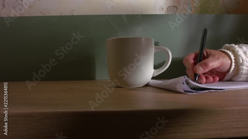 Woman writing with hot mug of coffee with writing pad in kitchen