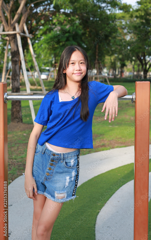 Fototapeta premium Portrait of young girl in blue shirt is leaning against the playground bars, while looking at camera.