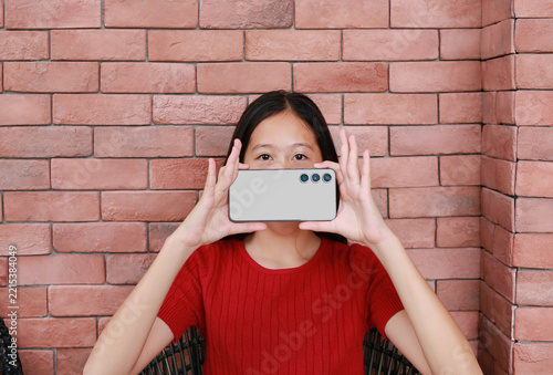 A young Asian girl in red shirt holds a smartphone horizontally in front of face, covering mouth and lower face, against red brick wall background.