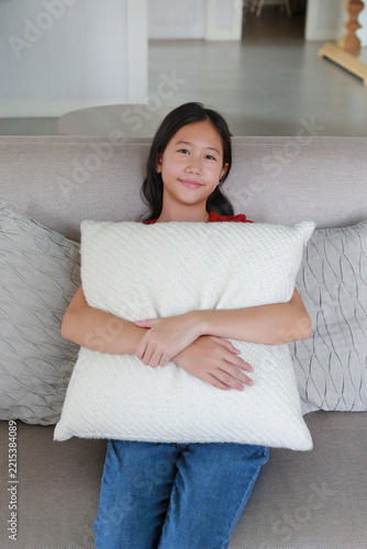 Young Asian girl sitting on a sofa and hugging a white cushion at home.