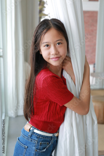 Young Asian girl standing by the window and holding a white curtain at home.