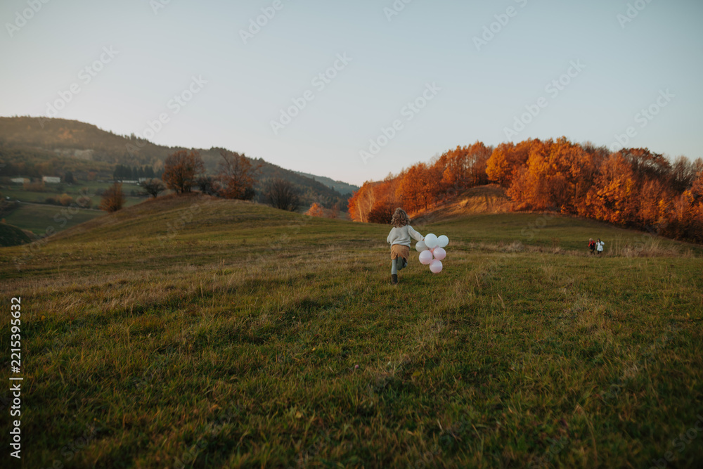 Naklejka premium Happy girl running with outstretched arms and pink and white balloons across a green autumn meadow.