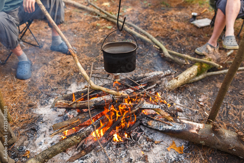 Metal pot hanging over campfire with burning logs surrounded by tourists sitting around