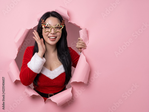 Surprised asian woman in red santa outfit peeking through torn pink paper hole