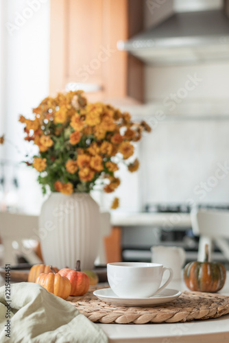 White tea cup with small pumpkins and orange flowers creates a relaxed autumn tea scene in a bright home kitchen. Vertical format.
