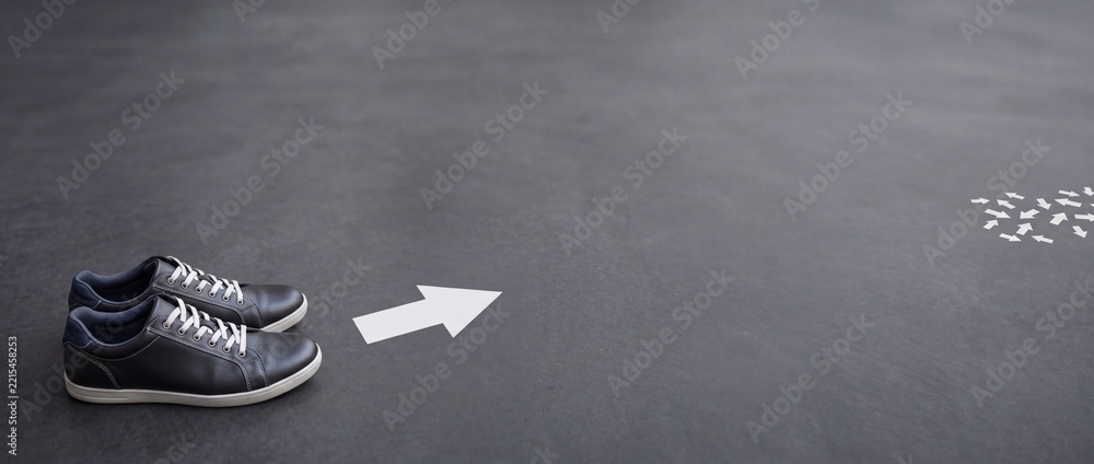 Fototapeta premium Pair of dark casual sneakers on a grey textured background with a white arrow pointing right towards a cluster of small arrows, representing choice, decision making, leadership, and direction.