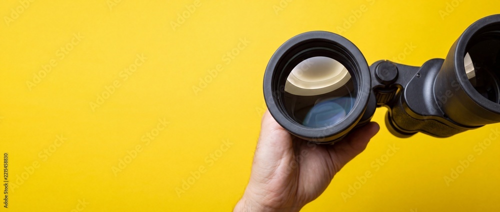 Fototapeta premium A hand holding black binoculars against a vibrant yellow background, symbolizing concepts of vision, future discovery, business strategy, looking for opportunities, and perspective shift.