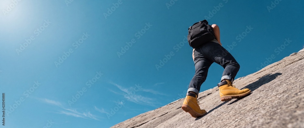 Naklejka premium Low angle view of a person with a backpack climbing a rocky mountain slope against a clear blue sky, capturing the spirit of outdoor adventure, determination, and achieving goals.