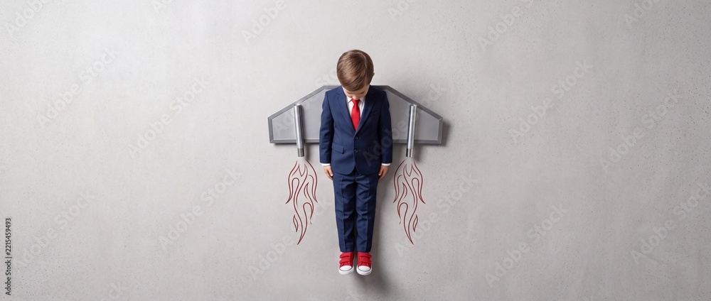 Fototapeta premium Young Boy in Business Suit Wearing Toy Jetpack Imagining Success and Future Career Growth, Creative Concept of Ambition and Childhood Dreams with Perspective Shift Top Down View