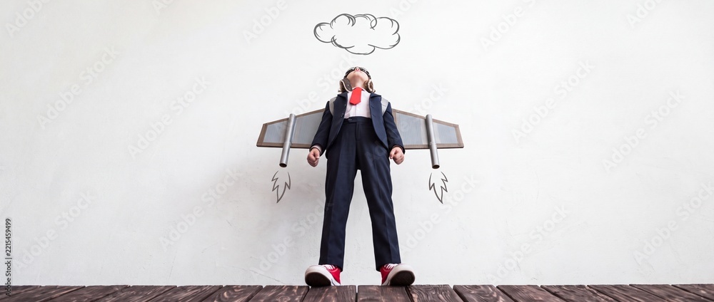 Fototapeta premium Ambitious young child in a formal business suit wearing aviator goggles and cardboard jetpack wings, looking up at hand-drawn clouds from a low angle perspective, symbolizing future career goals.