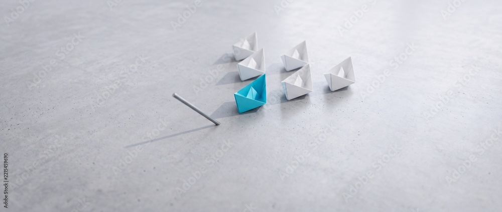 Fototapeta premium Blue paper boat leading a group of white origami boats on a grey stone surface, representing leadership, vision, and a shift in perspective for business success and strategic innovation.