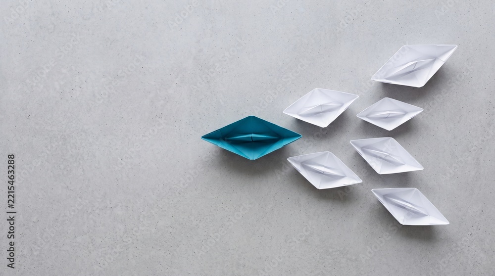 Fototapeta premium Top view of a unique blue paper boat leading a fleet of white boats on a grey textured background, conceptualizing leadership, business direction, teamwork, and standing out from the crowd.