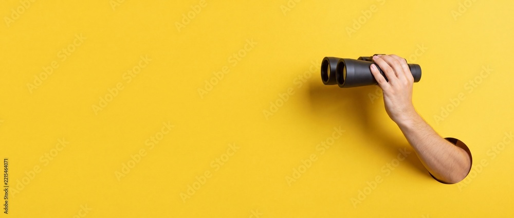 Fototapeta premium Hand holding black binoculars appearing through a round hole in a vibrant yellow background with wide copy space, representing discovery, business vision, and looking for new opportunities.