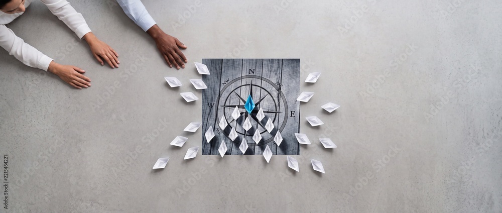 Fototapeta premium Business Leadership and Strategic Direction Concept Featuring a Blue Paper Boat Leading a Fleet of White Boats Over a Compass Graphic with Diverse People's Hands in a Wide Top View