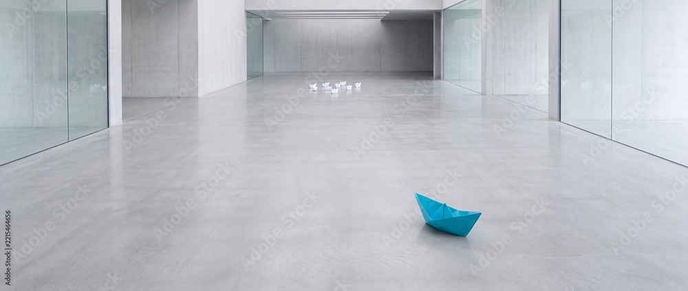 Fototapeta premium A single blue origami paper boat standing out from a group of white boats in a large minimalist modern architectural hall with concrete floors, representing leadership and individuality.