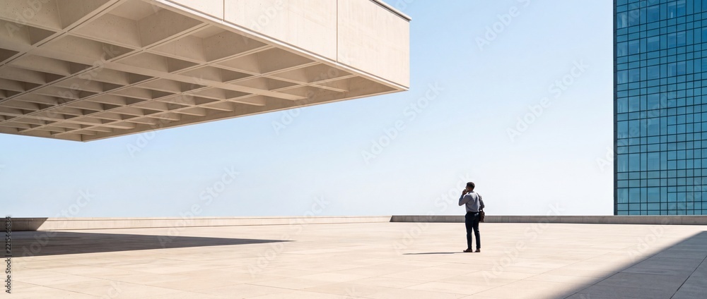 Fototapeta premium Professional businessman standing on a spacious outdoor terrace featuring minimalist brutalist architecture and a glass office building against a clear blue sky, symbolizing corporate vision.