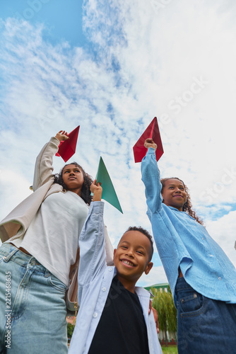 Sisters raising paper airplanes with younger brother in park with excited smiles. Creative learning, craft supplies, afterschool programs concept.