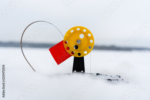 A yellow and red ice fishing tip-up stands ready in snow, acting as a reliable fishing flag. This functional fishing gear waits patiently on the frozen lake, partially covered by winter drifts.