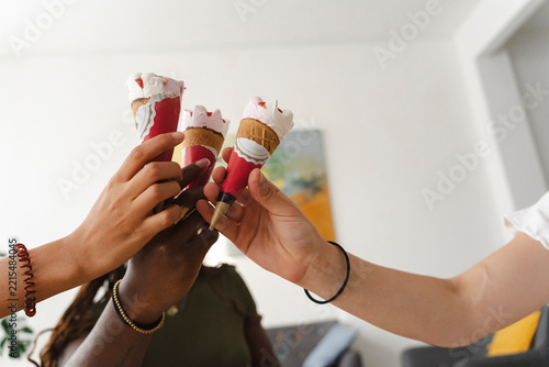 Three diverse friends hold their ice cream cones together in a celebratory toast indoors. They enjoy sharing a sweet treat during a casual gathering at home.