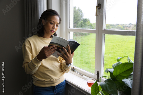 African woman standing by multi-pane window reading paperback book with glass and plant