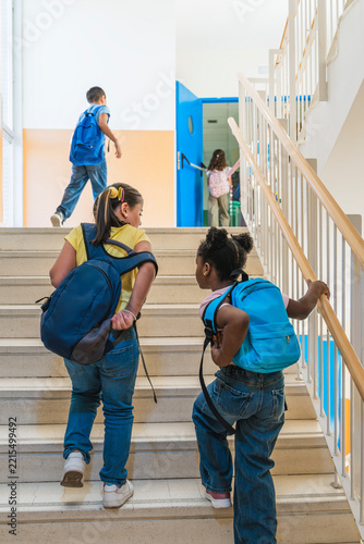 Diverse elementary school children with backpacks walking up stairs in a bright school building, heading to their classrooms