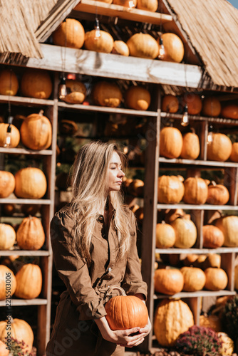 Woman holding a pumpkin at an outdoor autumn farm market surrounded by wooden shelves filled with pumpkins, warm sunlight, and seasonal harvest decorations