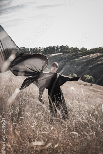 Young woman raises sweeping black textile overhead in dry field with rebellious energy. Defiance, self-expression, creative liberation concept