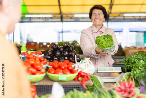 Smiling elderly female seller telling prices for fresh vegetables at local bazaar
