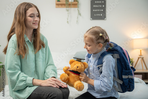 Happy little girl holding teddy bear and looking at her mother with a smile. Cheerful elementary schoolgirl wearing backpack holding graduation teddy bear and smiling at supportive mother in bedroom.