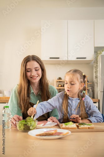 Happy little girl talking excitedly to mother while sitting at kitchen table with food. Cheerful daughter sharing funny moment with smiling mother during healthy meal preparation in home kitchen.