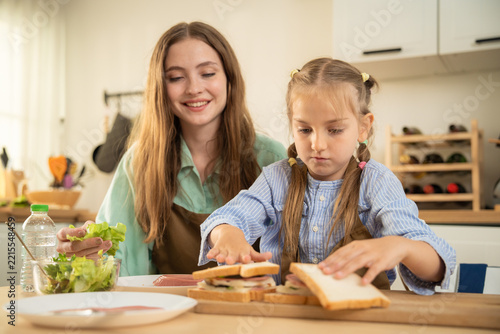 Happy little girl talking excitedly to mother while sitting at kitchen table with food. Cheerful daughter sharing funny moment with smiling mother during healthy meal preparation in home kitchen.