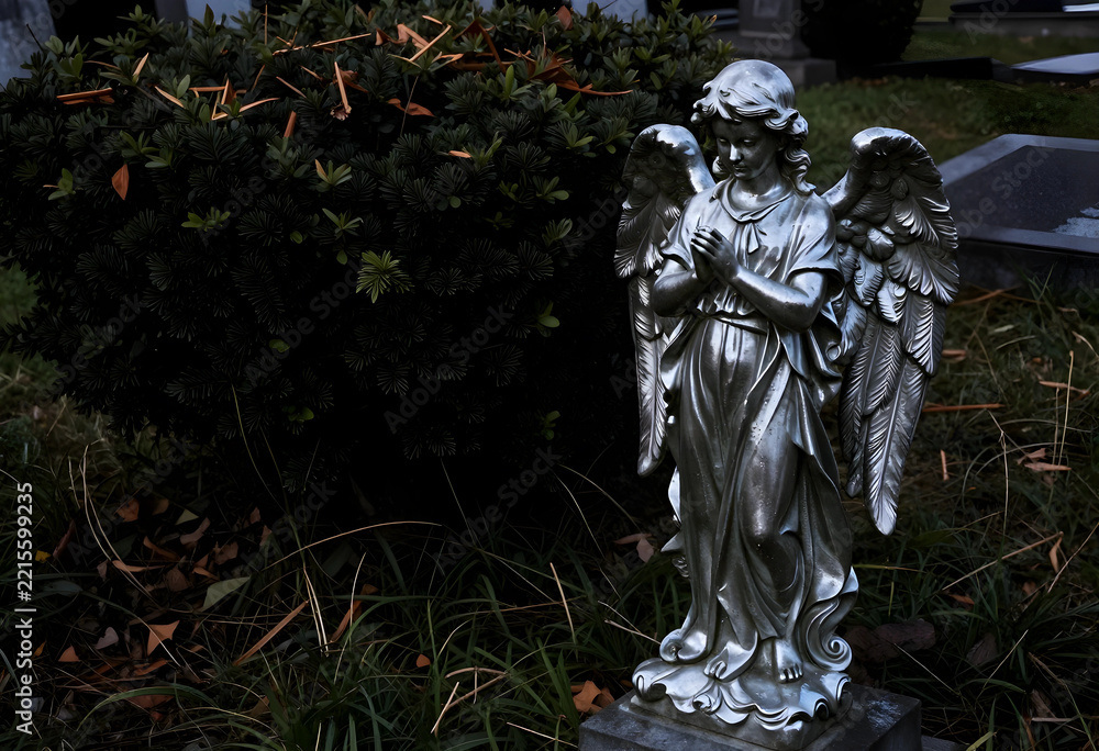 Fototapeta premium Grief and mourning concept, silver angel statue praying at a cemetery grave, religious memorial and funeral tribute in a quiet garden setting