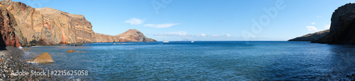 Panoramic view of a rocky volcanic bay with clear blue ocean, rugged cliffs and anchored sailboats under a bright sky in Madeira, Portugal