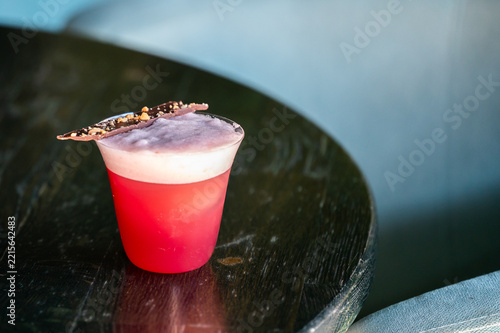 A vibrant red cocktail with a frothy foam layer sits on a dark wooden table, garnished with a piece of chocolate bark sprinkled with nuts.